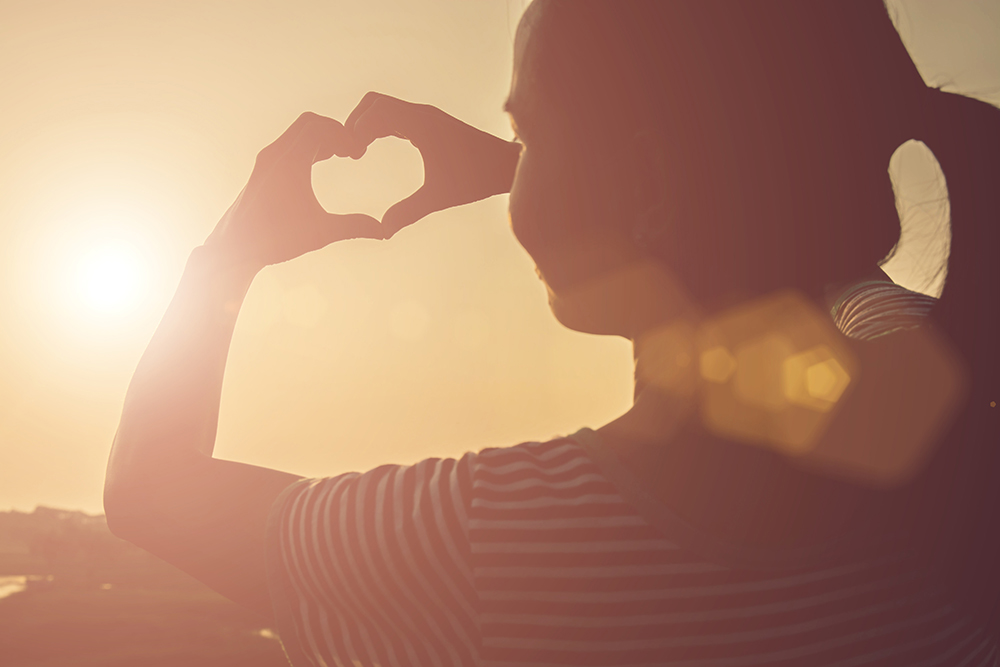 Young woman hands shaped heart against beautiful sunset